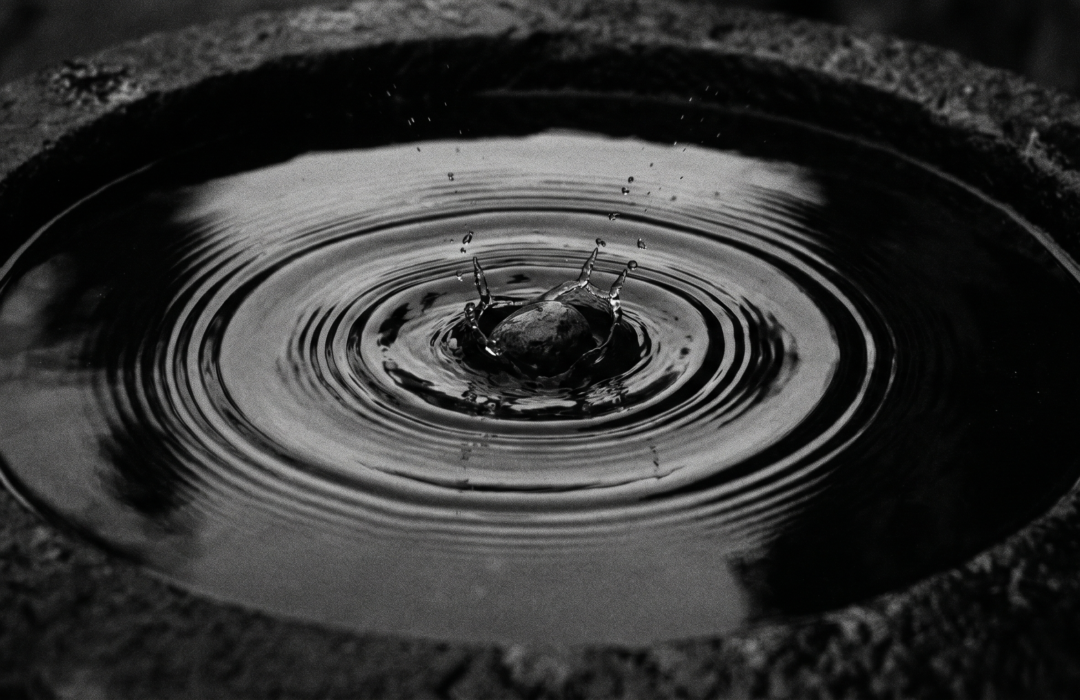 Fotografía en blanco y negro y clave baja de una pequeña piedra cayendo en el centro de un contenedor de piedra con agua, capturando el impacto exacto y creando ondas concéntricas perfectas sobre la superficie tersa. Estilo analógico con grano fino y alto contraste.