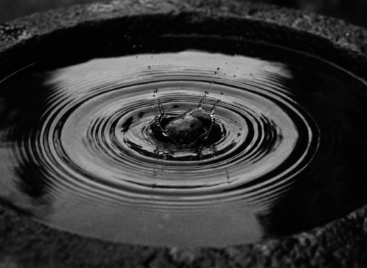 Fotografía en blanco y negro y clave baja de una pequeña piedra cayendo en el centro de un contenedor de piedra con agua, capturando el impacto exacto y creando ondas concéntricas perfectas sobre la superficie tersa. Estilo analógico con grano fino y alto contraste.